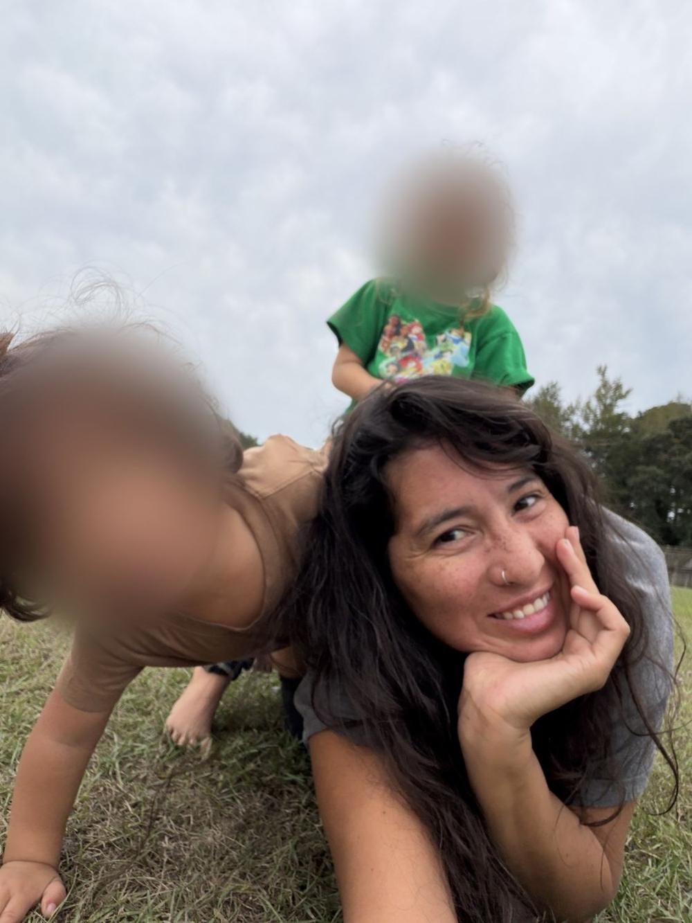 Paola Andrea lying in the grass, smiling, with her two daughters climbing on her back.